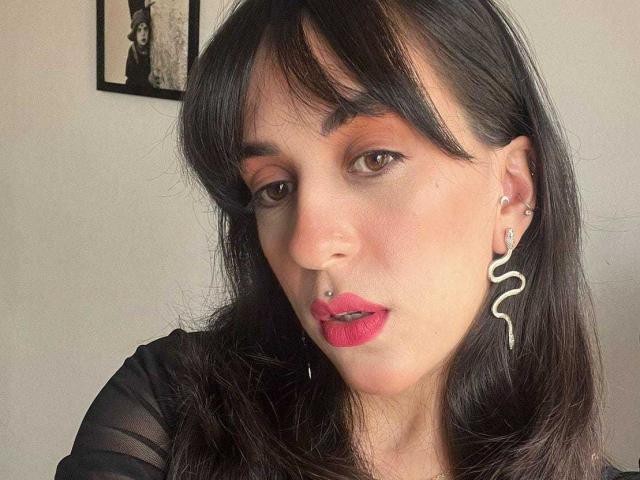 LunaStella has dark brown hair, red lipstick, and a nose ring. She's wearing a snake-shaped earring and a sheer black top. Her makeup includes pink eyeshadow.