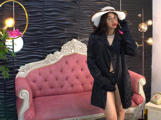 VictoriaCorins with dark hair, wearing a black coat, black gloves, and white hat, showing her bare ass, standing in front of a pink tufted sofa, holding her hat.