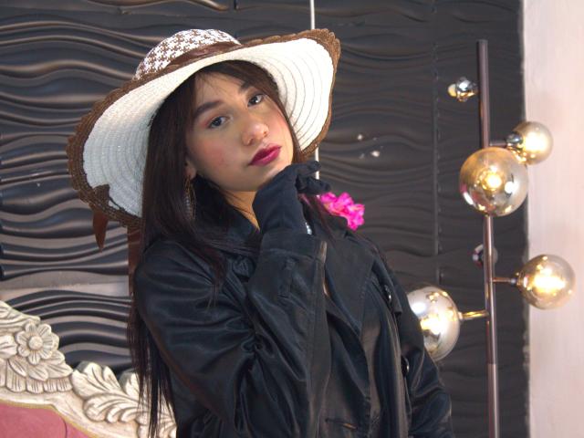 VictoriaCorins with long black hair, wearing a white sunhat and black jacket, touching her chin, showing pink lipstick and subtle blush, standing next to shiny metal balls.