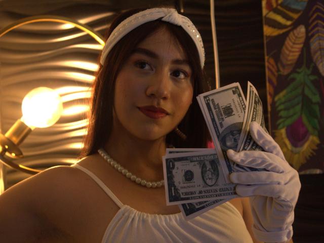 VictoriaCorins with brown hair, wearing white dress, pearl necklace, white headband, white gloves, holding a bunch of dollar bills, smiling, red lipstick, soft lighting.
