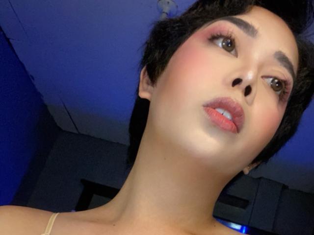TransFeminineSue tiene el cabello corto y negro, rubor rosa y lápiz labial rosa, con labios rosados ligeramente separados, mirando hacia la cámara, mostrando su rostro liso y de piel clara..