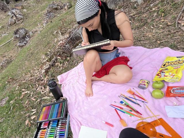 DeisyWest with black and white striped headscarf, black sleeveless top, and red skirt, sitting on pink blanket, drawing with colored pencils, surrounded by art supplies, apples, and snacks.