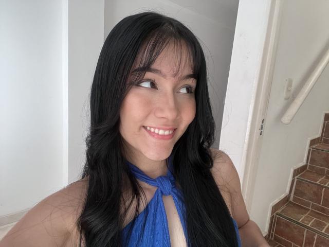 DeisyWest with long black hair, smiling, wearing a blue halter top that reveals her cleavage, standing near a stairway. She's looking to the side with cute freckles.