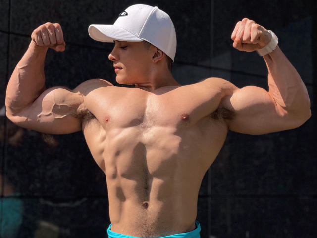 Muscular MaximoVega with flexed biceps, white cap, tan skin, and blue shorts, showing defined abs and pectorals, wearing a white wristband.