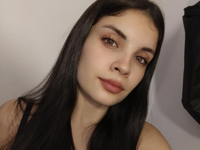 AriannaGray has long black hair, brown eyes, and pinkish lips with a slight smile. She's wearing a black top, and her skin is pale and smooth.