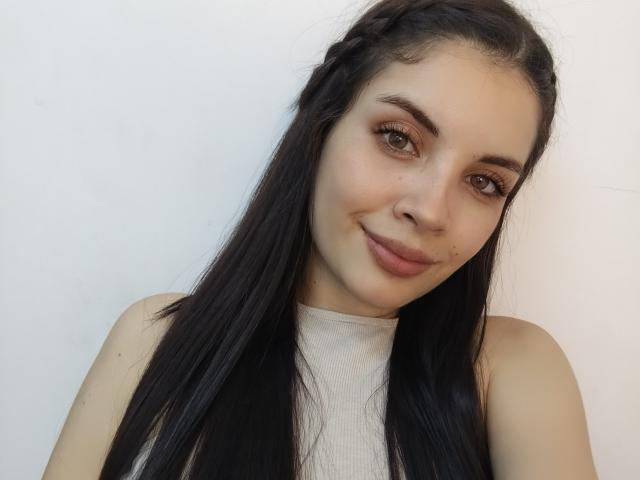 AriannaGray has long black hair, brown eyes, and a small smile with light makeup, wearing a white sleeveless top, showing bare shoulders. She has a cute, sexy face.