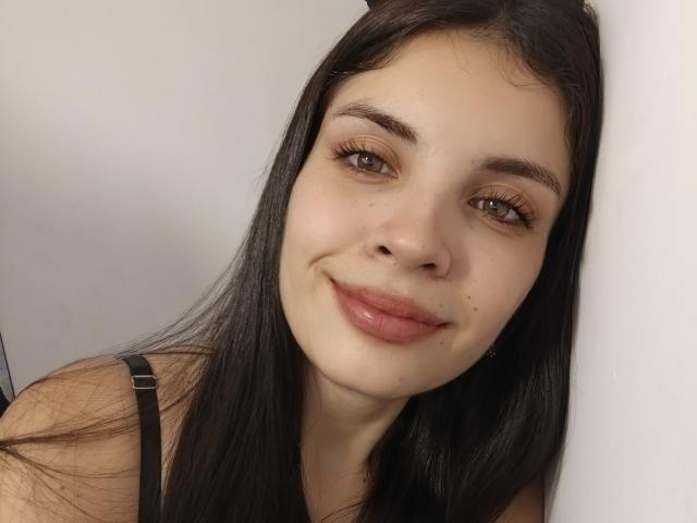 AriannaGray has fair skin, brown eyes, and long black hair. She's smiling with pinkish lips, wearing a black strap top, and has a small mole above her right lip.