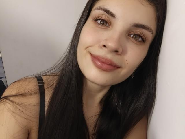 AriannaGray has long black hair, brown eyes, and pink lips. She's smiling slightly with a small mole above her right lip. She's wearing a black strap.