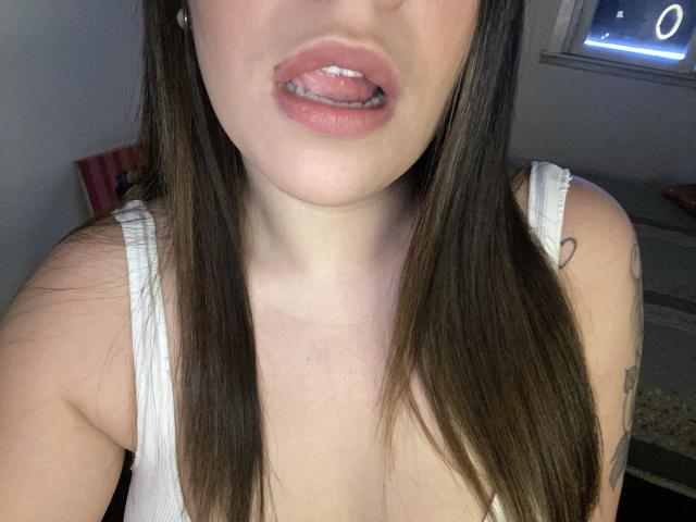 AlexaLip's face, showing her pink tongue slightly out, with brown straight hair, white tank top, and a small heart tattoo on her right arm. Boobs, pale skin, pink lips.