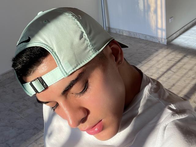 MaximoVega with eyes closed, wearing a light green baseball cap, white shirt, and small earrings, sunlit skin, black eyebrows, short dark hair, pink lips, marble floor, shadows.