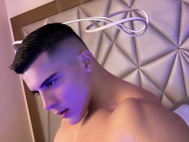 MaximoVega, shirtless, with short black hair, a diamond earring, and a purple light on his face, looking down, has a sexy, muscular, and attractive appearance.