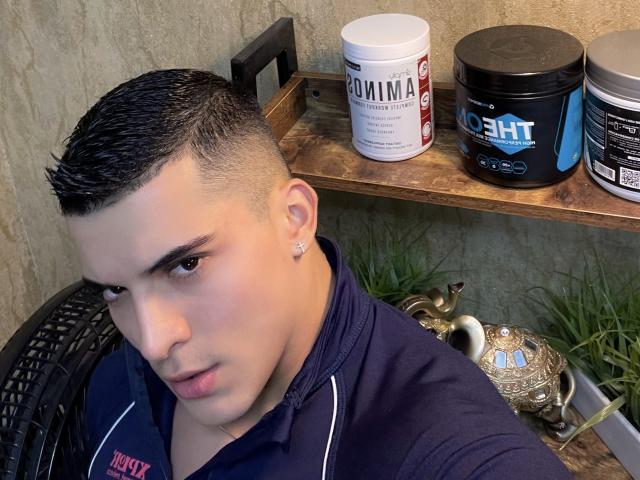 MaximoVega with short, dark, slicked-back hair, wearing a navy polo, has a stud earring, and is looking at the camera with a slight frown, sitting near supplements.