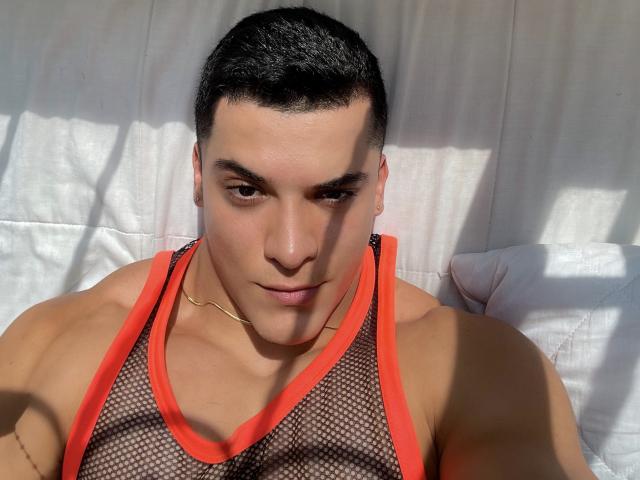 MaximoVega with short black hair, wearing a red mesh tank top, gold chain, and serious expression, sunlight casting shadows on his muscular, tan-skinned face and chest.