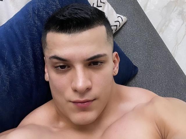 MaximoVega's chiseled face with short, dark hair, small ear piercings, and a neutral expression, lying shirtless with visible, toned shoulders. His brown eyes are slightly downcast.