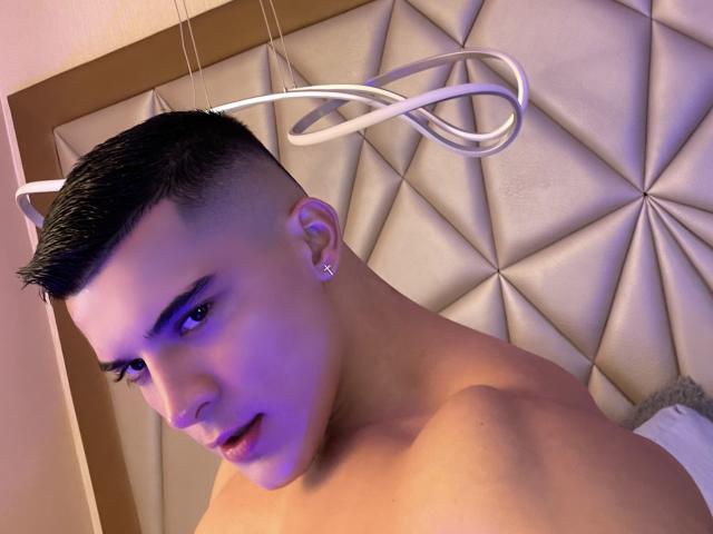 MaximoVega has short black hair, a diamond earring, and a purple-blue light on his face, shirtless with a neutral expression, and a halo-like light above his head.