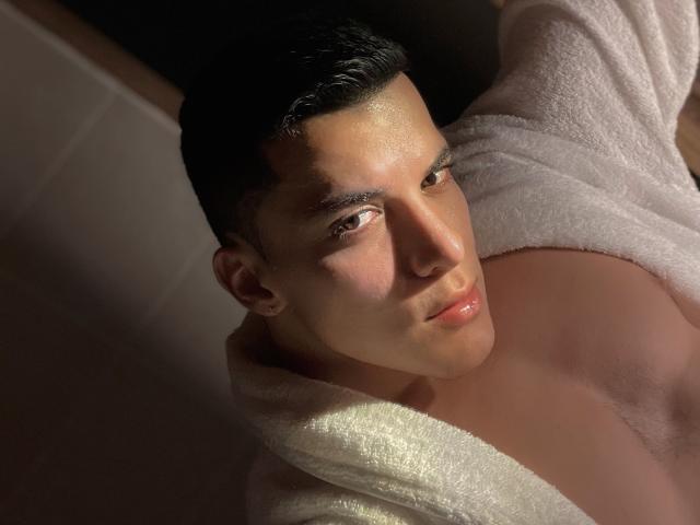 MaximoVega with short black hair, glistening skin, and a white towel, looking up with soft pink lips, slightly parted, in dim lighting, showing a strong, attractive face.