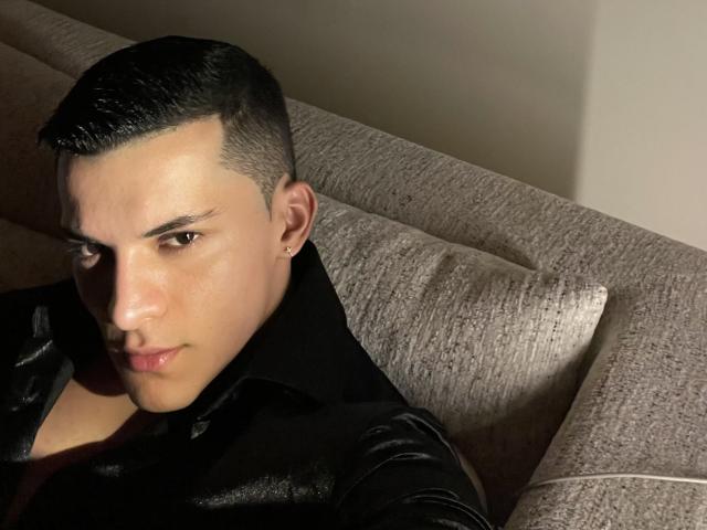 MaximoVega with short black hair, leather shirt, earring, and serious expression, lying on a gray textured couch, shadow on his face, slightly parted lips.