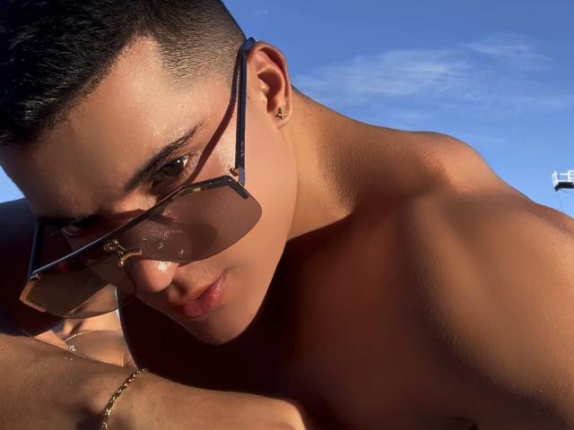 MaximoVega with tan skin, short dark hair, gold earring, and sunglasses, leaning forward, shirtless, sunlight on his chest, blue sky in background, wearing a gold bracelet.