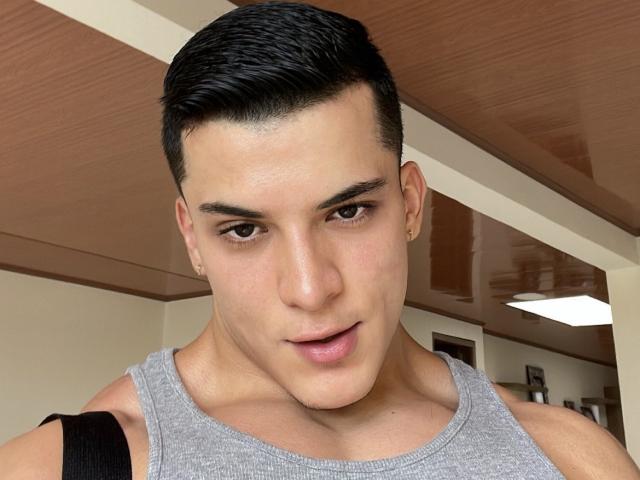 MaximoVega with short black hair, gray tank top, small earrings, and a black strap, looking down with slightly parted pink lips. His skin is tan and he's handsome.