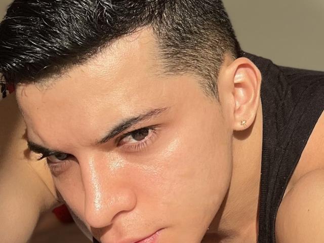 MaximoVega's sweaty face with dark, short, wet hair, brown eyes, and a small stud earring in his left ear, wearing a black tank top, looking intense and focused.