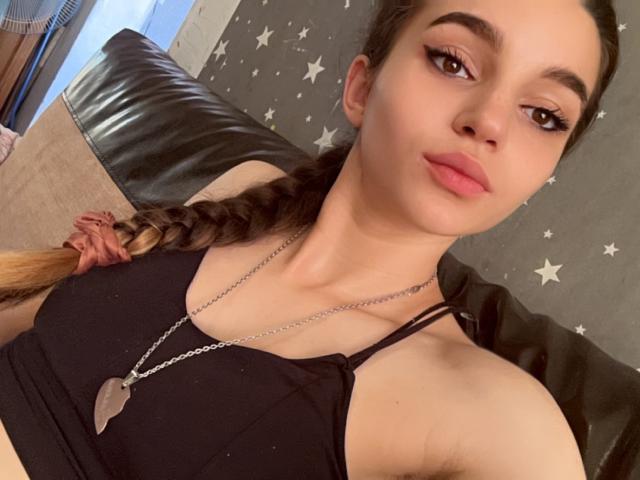 Tiffany Campbell, com cabelo trançado, camiseta preta e colar, mostrando os seios e lábios rosados e curvados, olhando para a câmera com os olhos semicerrados..