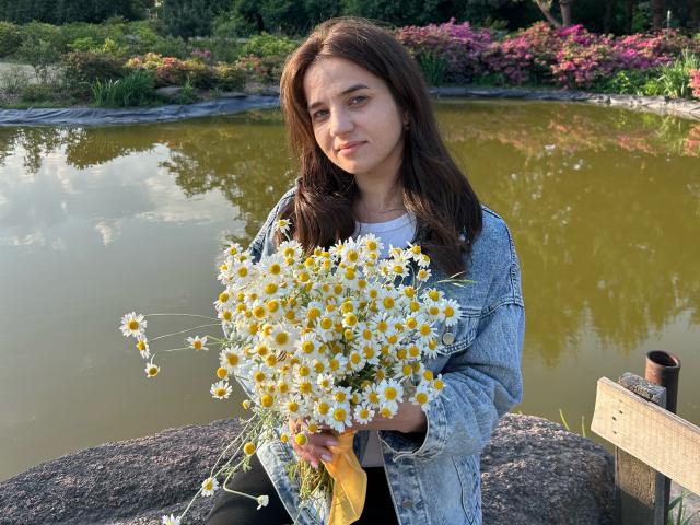 JacquelynCelie met bruin haar, die een boeket witte margrietjes met gele centra vasthoudt, gekleed in een denimjas, staand bij een groenachtig meer met een houten omheining..