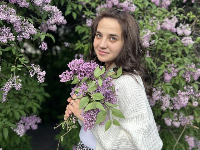 JacquelynCelie, avec ses cheveux bruns ondulés, tenant un bouquet de lilas violets, portant un pull blanc en tricot, souriant légèrement, debout dans un jardin luxuriant de couleurs vertes et violettes..