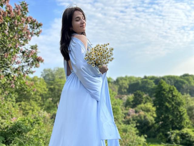 JacquelynCelie vestida de branco, segurando um buquê de pequenas flores amarelas, em um ambiente externo com árvores verdes e um céu azul com nuvens esparsas ao fundo..