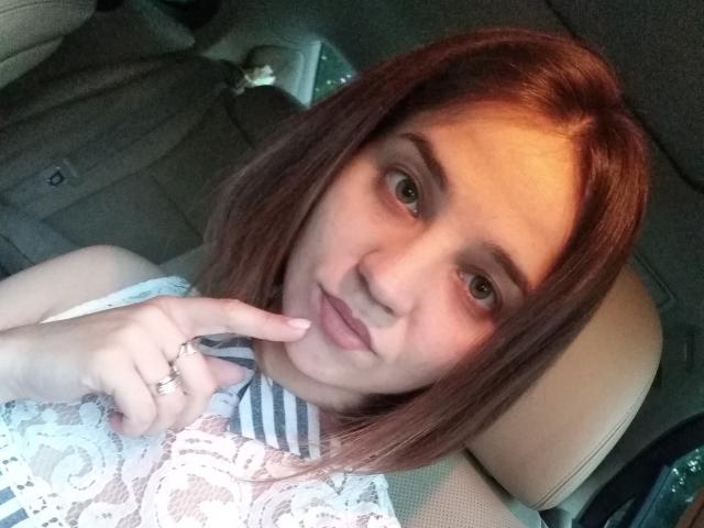 JacquelynCelie with brown hair, green eyes, and a ring on her right index finger, touching her lips with her finger, wearing a white lace dress, in a car.