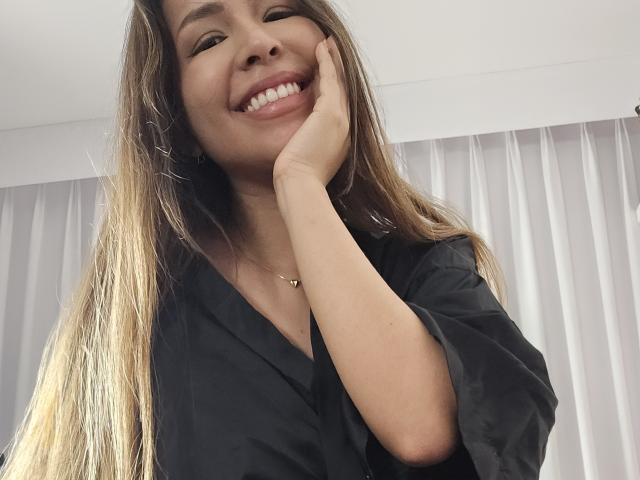 MeganWisse with long blonde hair, smiling, hand on cheek, wearing a black shirt, light skin, cute, smiling teeth, subtle necklace, upper body, casual photo.