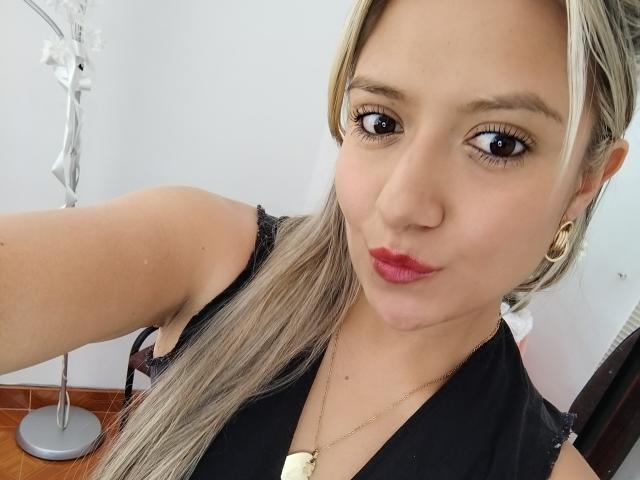 ValeriaHunter69 with light brown skin, blonde ponytail, red lipstick, black sleeveless top, gold necklace, small gold earrings, and a cute smile, taking a selfie in a bright room.