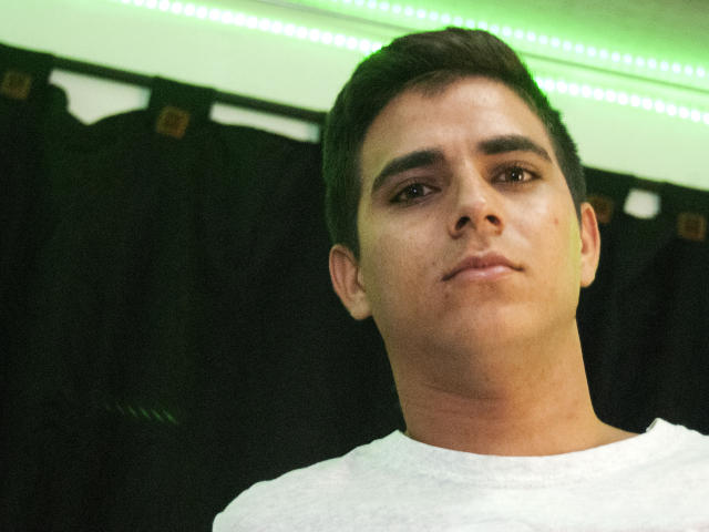 SantosMontenegro with short dark hair, slight beard, and white shirt, looking slightly up with a neutral expression, green LED strip in background, slightly blurry, realistic.