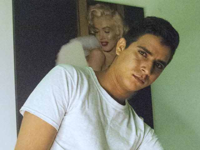 SantosMontenegro with short black hair, white t-shirt, serious expression, in front of a picture of Marilyn Monroe's bare ass and face in the background.