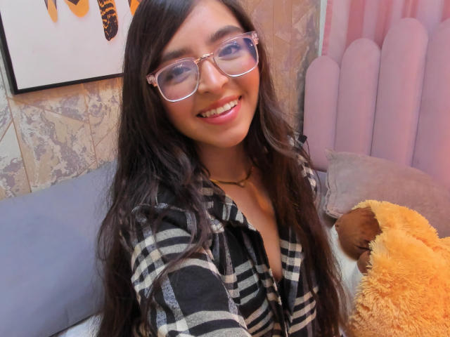 Eva McAllister, com cabelos longos e escuros, óculos, sorrindo, vestindo uma camisa xadrez, segurando um brinquedo marrom e branco fofo, com uma cabeceira rosa ao fundo..