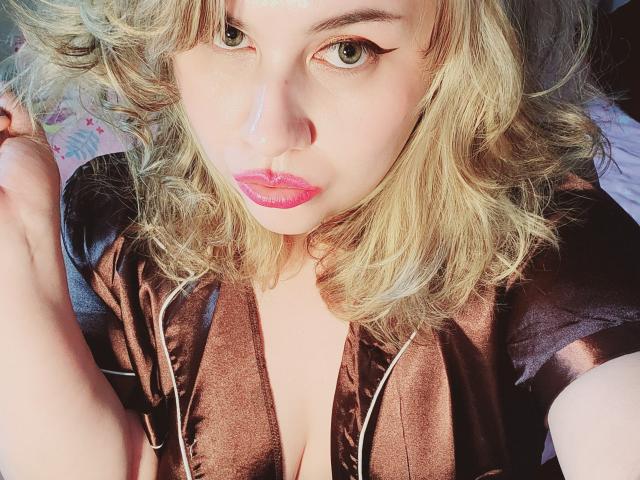 Akkanee with blonde curly hair, wearing a brown satin robe, has pink lipstick, and black eyeliner, looking up with green eyes, and holding her hair on the left.