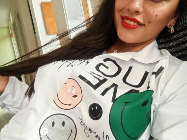 MadissonFontaine with long brown hair, red lipstick, and freckles, wearing a white shirt with cute face prints, holding her hair, smiling, showing her tits, in a chair.
