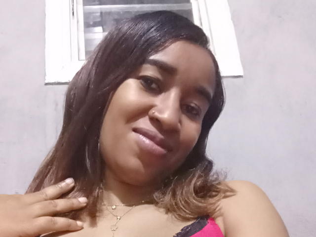 FloJolie with medium brown skin, dark brown hair, and a nose ring, smiling slightly, wearing a red and black lace top, touching her chest with her right hand.