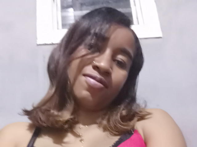 FloJolie has medium brown skin, shoulder-length wavy brown hair, and is wearing a pink and black bra, looking down with a slight smile. She has a gold necklace.