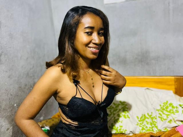 FloJolie with medium brown skin and black lace bra, showing her tits, smiling, hand on chest, brown wavy hair, gray wall background, wooden bed with green patterned pillow.