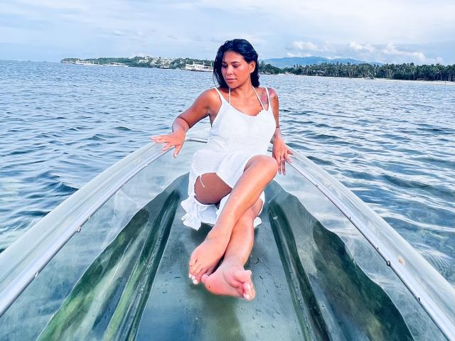 SexyKaila in a white dress, sitting in a glass boat, legs crossed, hands on edges, water in background, bare feet, dark hair, blue eyes, water droplets on boat.