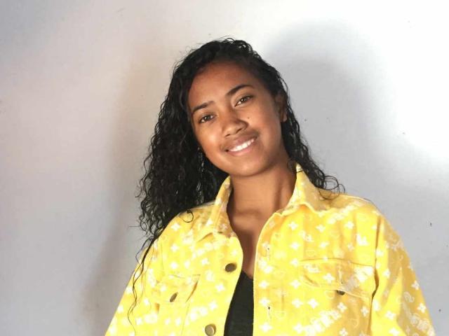 Enielah with curly black hair, smiling, wearing a yellow patterned shirt, and a black top, has brown eyes, and dark skin, standing against a white wall.