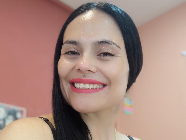 CharlotteCrofort has shiny skin, black straight hair, red lipstick, and a nose piercing. She's smiling with pink lips and a silver nose ring. Her tits are not visible.