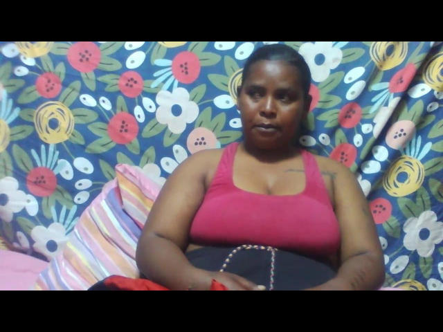 Latipha, a dark-skinned woman with a curvy build, wearing a pink tank top and black pants, sits with a red bracelet on her wrist. Her tits are visible.