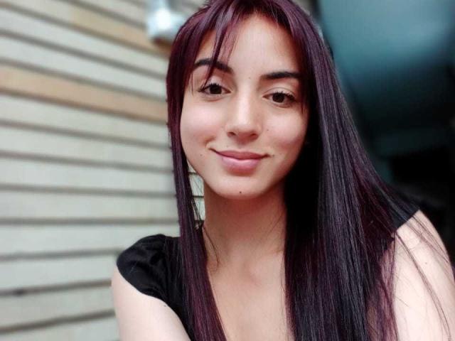 LourenJhonsson tiene el cabello largo, lacio y de color púrpura oscuro, piel de color marrón claro, y está sonriendo suavemente con ojos pequeños y marrones y una nariz pequeña y linda. Lleva una blusa negra..