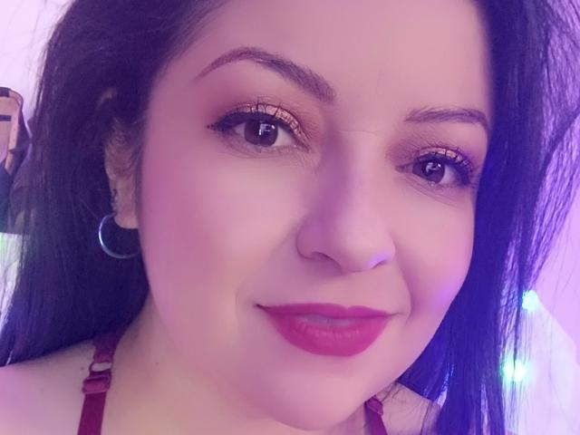 NatalyHami's face with purple-lit skin, black hair, red lipstick, shiny brown eyes, and a subtle smile. She's wearing a red strap, hoop earrings, and has a nose ring.