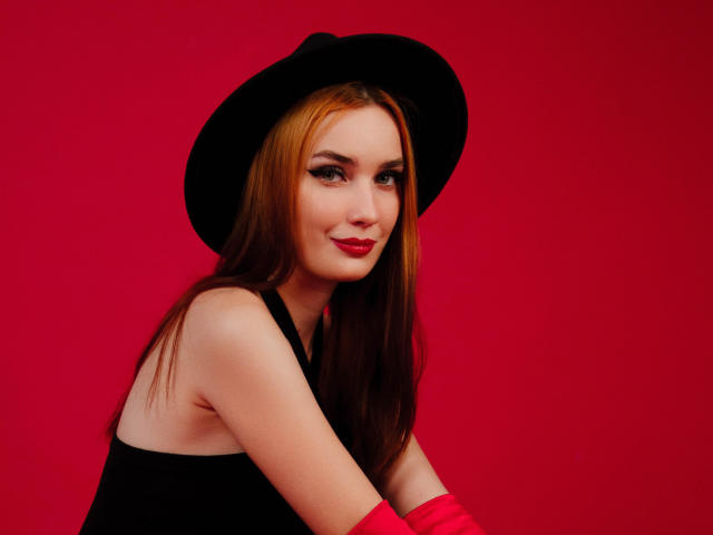 AlexisHott with red lipstick, black hat, and pink gloves, smiling, showing bare shoulders, and red background, with her tits slightly visible, looking sexy and confident.