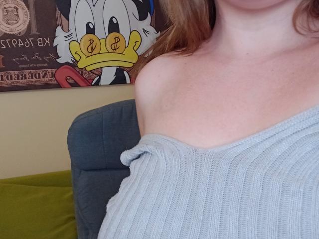 DiLaria's off-shoulder gray ribbed sweater shows her bare shoulders and upper chest. A Disney Duck character poster is in the background. Her red hair is partially visible.