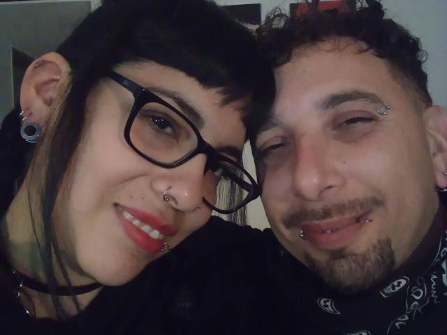 MarianneyRoger heeft zwarte brillen, neus piercings en rode lippenstift. De vriend van MarianneyRoger heeft krullend haar, een baard en piercings in zijn gezicht. Ze glimlachen allebei en leunen tegen elkaar aan..