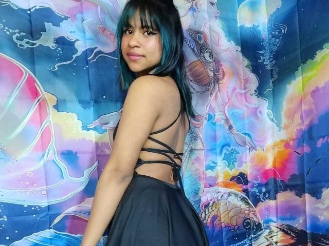 StarGirls69 with teal-dyed hair, black strappy dress, looking over shoulder, showing her back and ass, standing in front of colorful, psychedelic wall art.