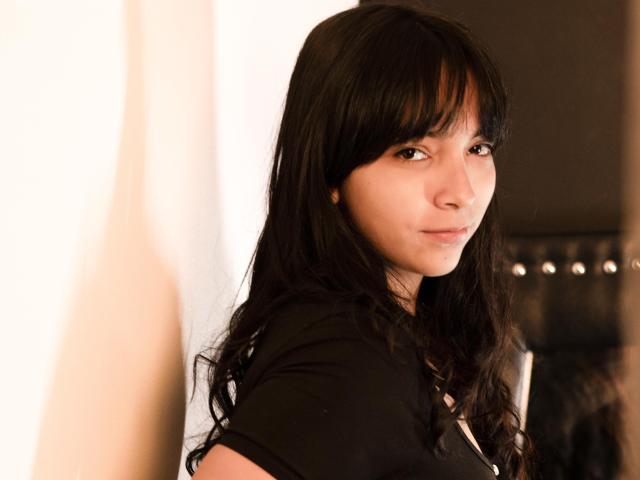 Nessa69s with long black hair, bangs, and a slight smile, wearing a black shirt, has a cute face, and small, cute nose, standing in a softly lit room.
